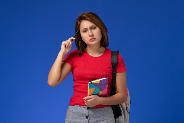 Vista frontal jovem aluna em camisa vermelha, usando mochila segurando o caderno com caneta sobre fundo azul claro.