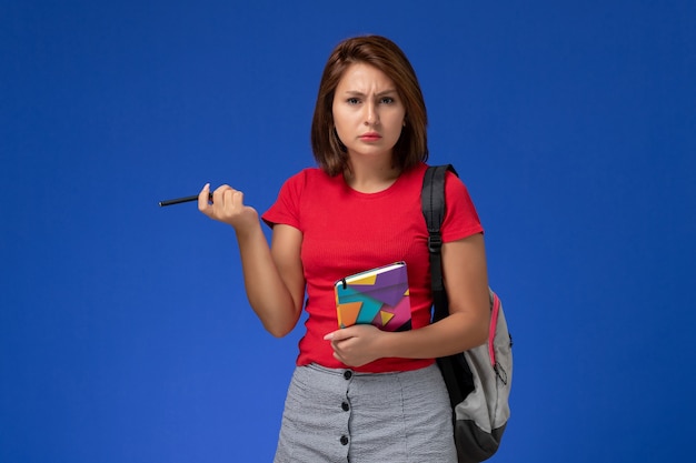 Vista frontal jovem aluna em camisa vermelha, usando mochila segurando o caderno sobre fundo azul claro.