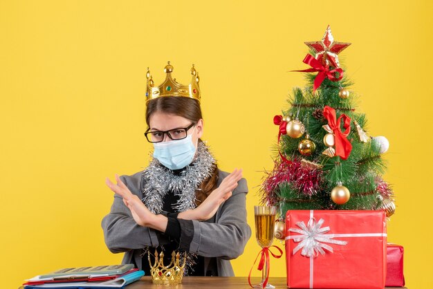 Vista frontal jovem com coroa usando máscara e óculos cruzando as mãos árvore de natal e coquetel de presentes