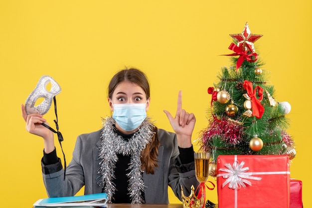 Vista frontal jovem com máscara médica sentada à mesa segurando máscara de baile de máscaras árvore de natal e coquetel de presentes