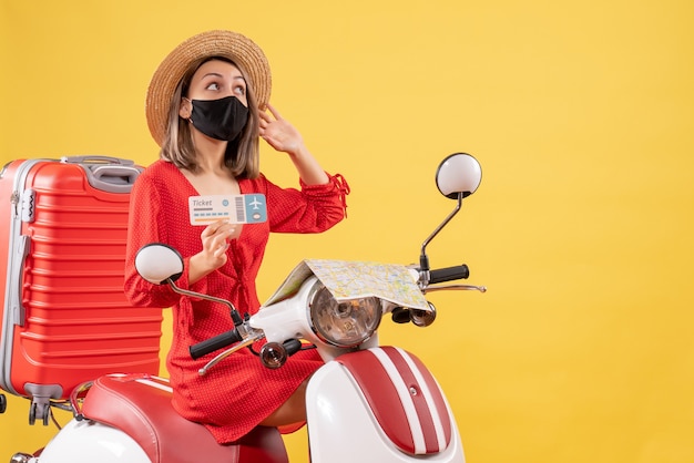 Vista frontal jovem com máscara preta em ciclomotor com mala vermelha segurando bilhete olhando para algo