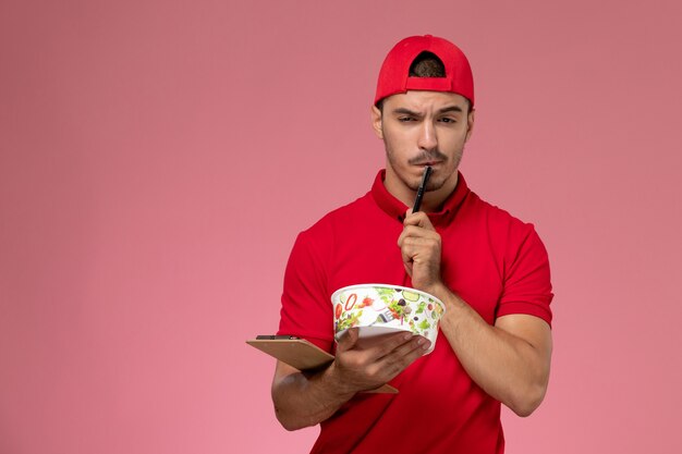 Vista frontal jovem correio masculino com capa uniforme vermelha segurando a tigela de entrega e o bloco de notas pensando na mesa-de-rosa.