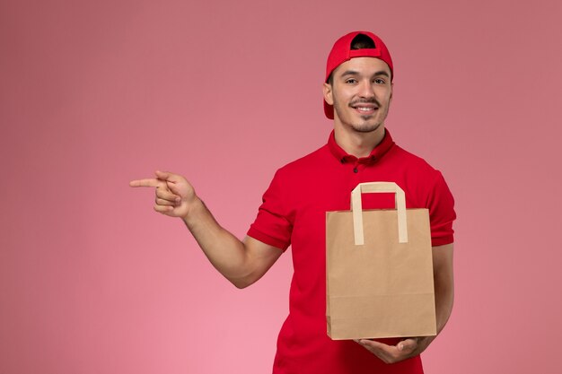 Vista frontal jovem correio masculino com capa uniforme vermelha, segurando o pacote de comida de papel no fundo rosa.