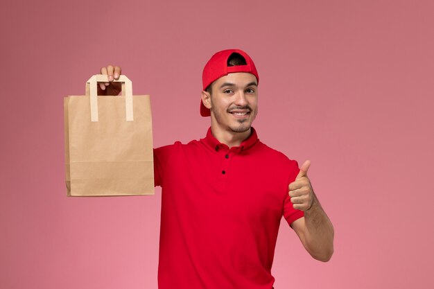 Vista frontal jovem correio masculino com capa uniforme vermelha, segurando o pacote de comida de papel, sorrindo sobre o fundo rosa claro.