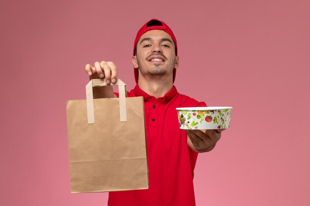 Vista frontal jovem correio masculino com capa uniforme vermelha, segurando o pacote de comida e a tigela no fundo rosa.