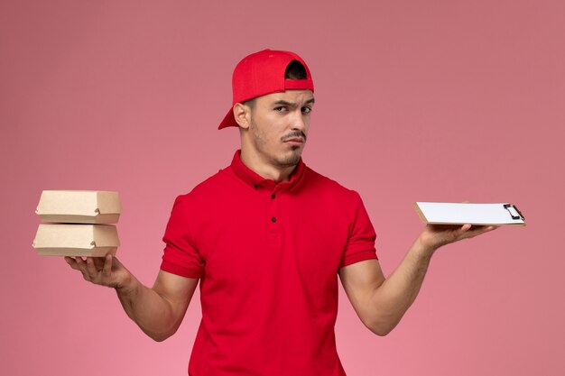 Vista frontal jovem correio masculino com capa uniforme vermelha segurando pequenos pacotes com comida e o bloco de notas sobre fundo rosa claro.