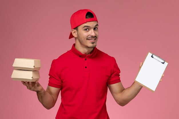 Vista frontal jovem correio masculino com capa uniforme vermelha segurando pequenos pacotes com comida e o bloco de notas sobre fundo rosa claro.