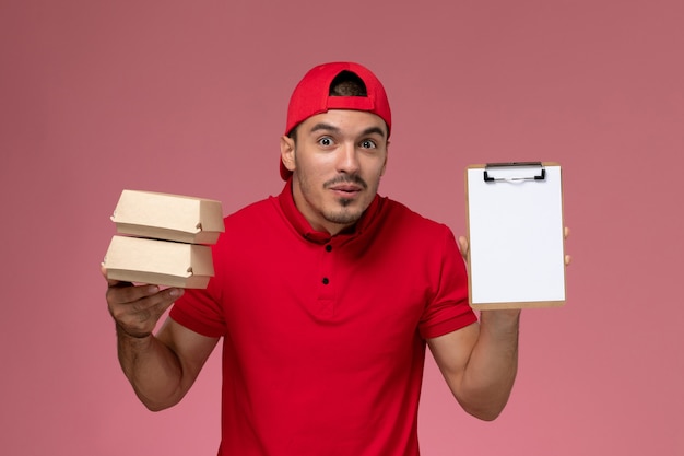 Vista frontal jovem correio masculino com capa uniforme vermelha segurando pequenos pacotes com comida e o bloco de notas sobre fundo rosa claro.