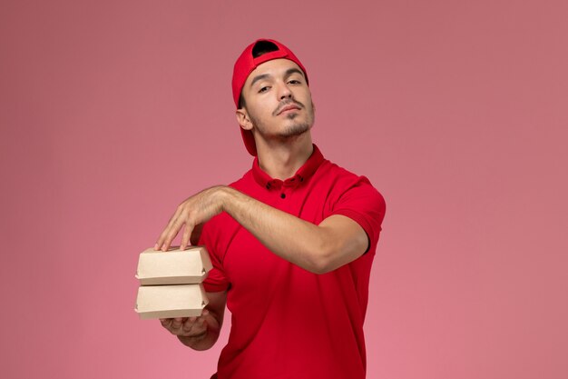 Vista frontal jovem correio masculino com capa uniforme vermelha segurando pequenos pacotes com comida no fundo rosa claro.