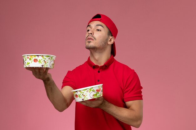 Vista frontal jovem correio masculino com capa uniforme vermelha segurando tigelas de entrega no fundo rosa claro.