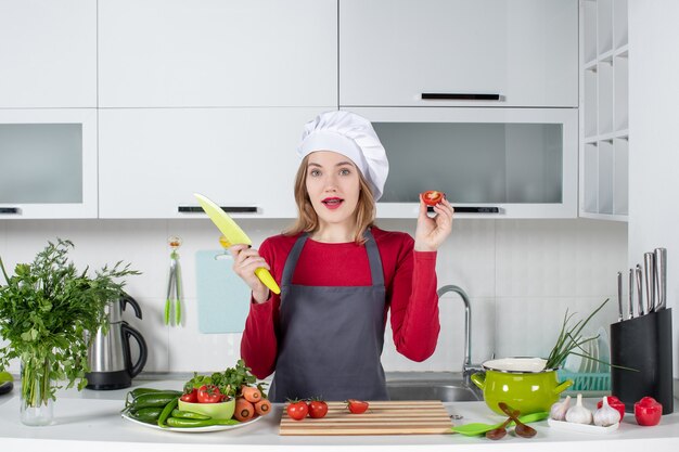 Vista frontal jovem cozinheira de avental segurando uma faca e um tomate