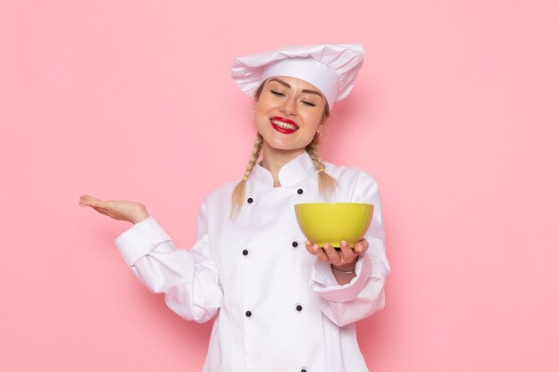 Vista frontal jovem cozinheira em terno branco, segurando uma placa verde com um sorriso no espaço rosa, cozinhar comida refeição