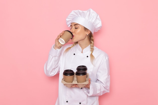 Foto grátis vista frontal jovem cozinheira em terno branco segurando xícaras de café de plástico no espaço rosa.