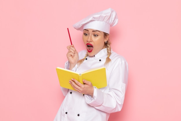 Vista frontal jovem cozinheira em traje branco de cozinheira escrevendo notas sobre o cozinheiro do espaço rosa