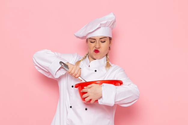 Vista frontal jovem cozinheira em traje branco misturando tigela vermelha no espaço rosa.