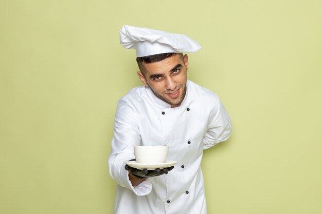 Vista frontal jovem cozinheiro em terno branco de cozinheiro usando luvas escuras e servindo café no verde