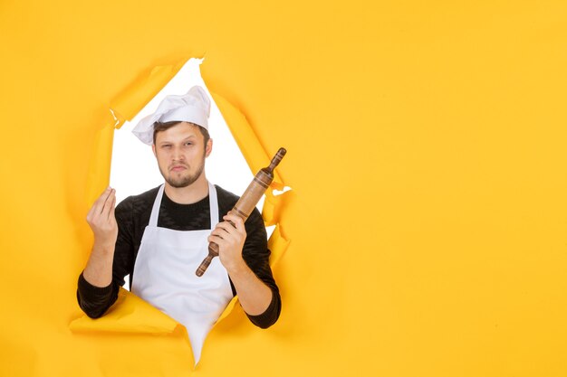 Vista frontal jovem cozinheiro masculino com capa branca segurando o rolo sobre fundo amarelo foto comida homem branco cozinha cozinha cores de trabalho