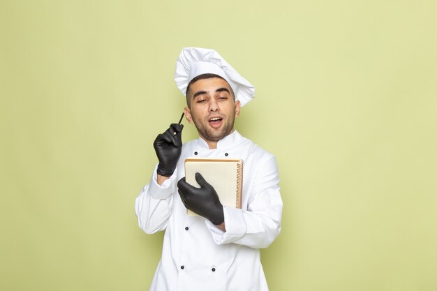 Vista frontal jovem cozinheiro masculino em terno branco de cozinheiro escrevendo notas com luvas no verde