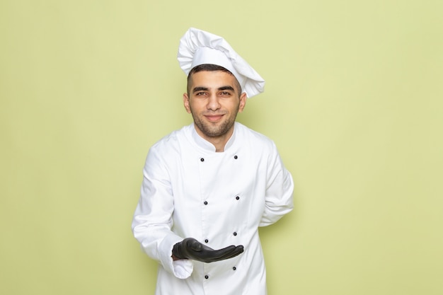 Vista frontal jovem cozinheiro masculino em terno branco de cozinheiro usando luvas pretas e dando boas-vindas no verde