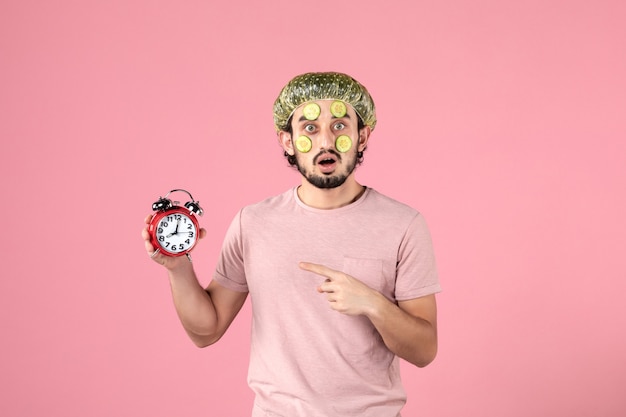 vista frontal jovem do sexo masculino com máscara no rosto segurando o relógio no fundo rosa