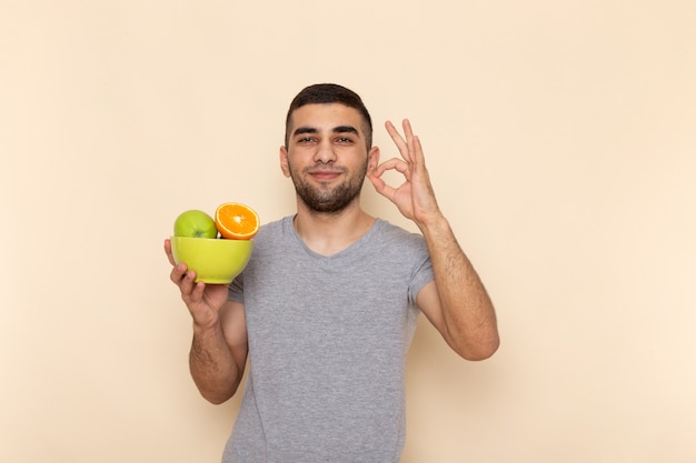 Vista frontal jovem do sexo masculino em camiseta cinza e jeans azul, sorrindo e segurando um prato com frutas em bege