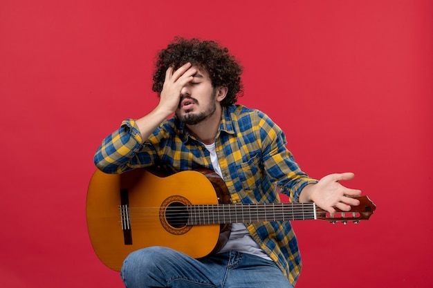 Vista frontal jovem do sexo masculino sentado com guitarra na parede vermelha música performance músico cor aplausos tocar concerto ao vivo