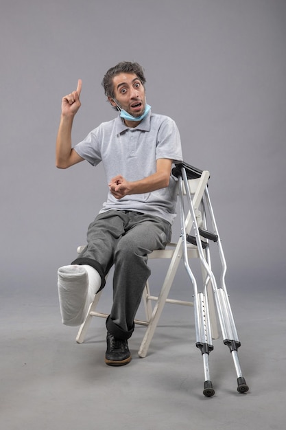 Vista frontal jovem do sexo masculino sentado com o pé quebrado e bandagem amarrada na parede cinza Dor no pé torção na perna acidente masculino