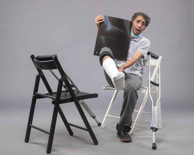 Vista frontal jovem do sexo masculino sentado com o pé quebrado e segurando um raio-x na parede cinza.