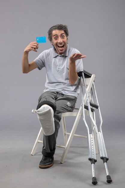 Vista frontal jovem do sexo masculino sentado com o pé quebrado segurando um cartão do banco azul na parede cinza Dor no pé quebrado torção acidente masculino