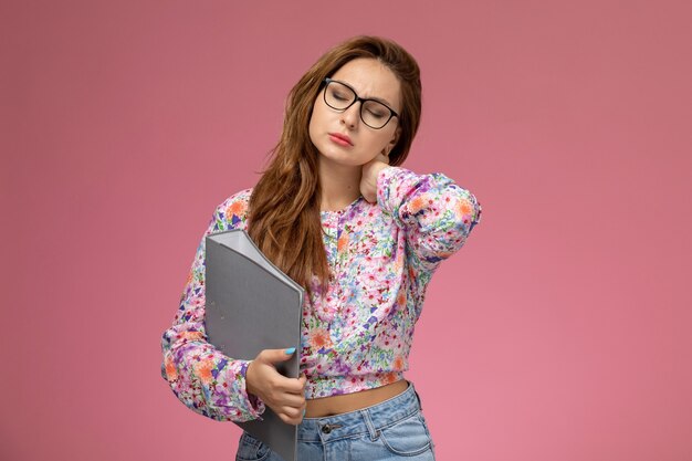 Vista frontal, jovem e bela mulher em uma camisa com design floral e jeans azul, com dor no pescoço no fundo rosa