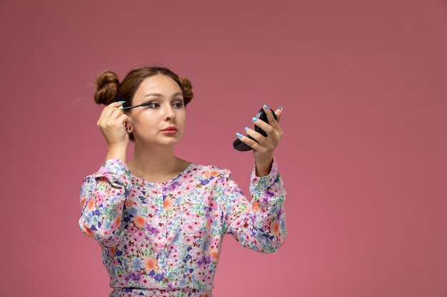 Vista frontal, jovem e linda mulher em uma camisa com design floral e jeans azul fazendo maquiagem no fundo rosa