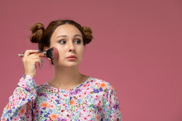 Vista frontal, jovem e linda mulher em uma camisa com design floral e jeans azul fazendo maquiagem no fundo rosa