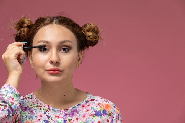 Vista frontal, jovem e linda mulher em uma camisa com design floral e jeans azul fazendo maquiagem no fundo rosa