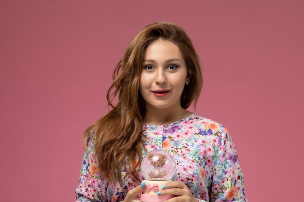 Vista frontal, jovem e linda mulher em uma camisa com design floral e jeans azul segurando um pequeno brinquedo de vidro sorrindo no fundo rosa