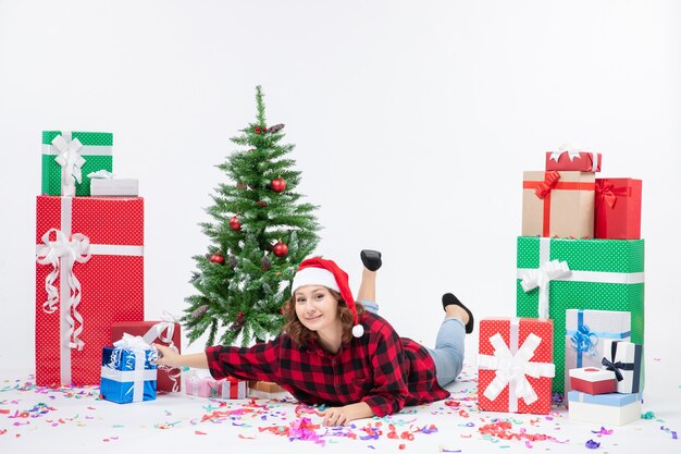 Vista frontal jovem fêmea deitada ao redor de presentes de natal e pequena árvore de férias no fundo branco mulher fria natal neve ano novo