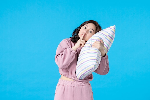 Vista frontal jovem feminina de pijama rosa segurando o travesseiro perto da cabeça na parede azul noite cor festa insônia cama descanso sonho mulher dormir