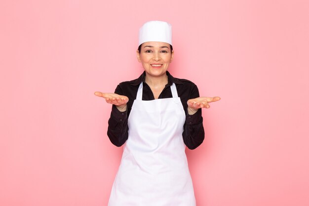 Vista frontal, jovem, femininas, cozinheiro, em, camisa preta, cozinheiro branco, capa, boné branco, posando, meio sorriso, encantado, sorrindo