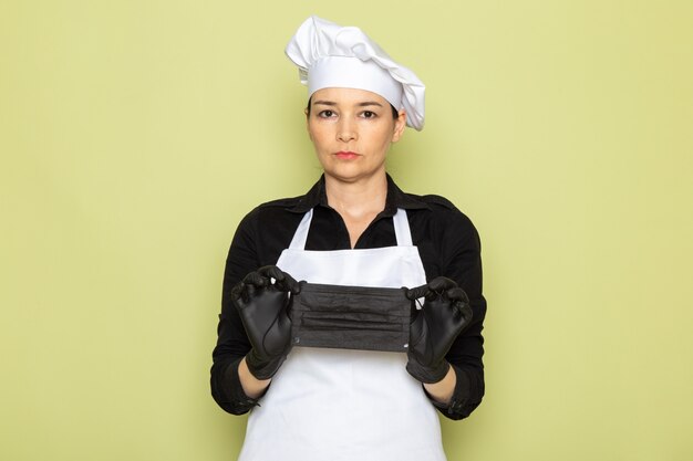 Vista frontal, jovem, femininas, cozinheiro, em, camisa preta, cozinheiro branco, capa, boné branco, posar luvas pretas, segurando, máscara preta, posando