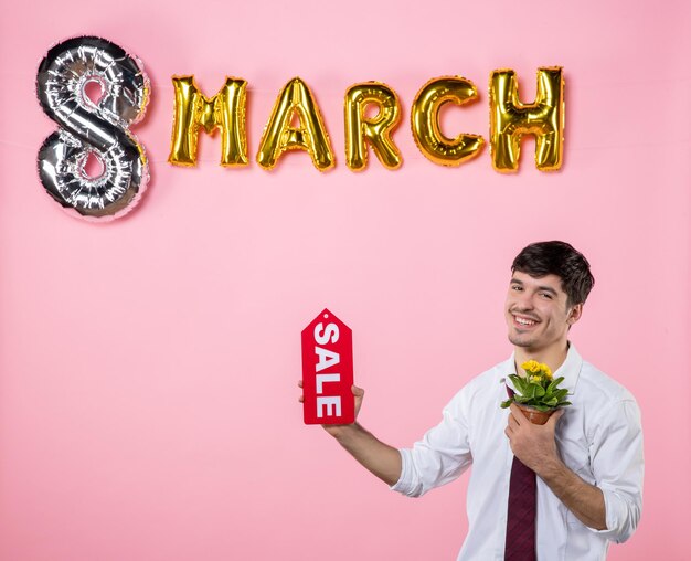 Vista frontal jovem homem segurando placa de identificação de venda vermelha com decoração de março em fundo rosa dinheiro compras homem dia das mulheres igualdade cor de feriado feminino