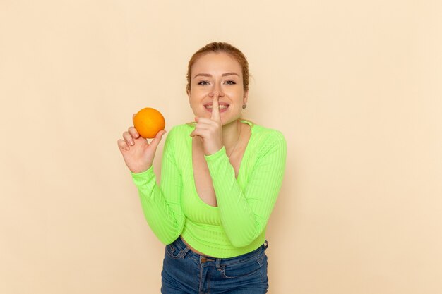 Vista frontal jovem linda mulher com camisa verde segurando laranja fresca sorrindo na parede creme fruta modelo mulher suave