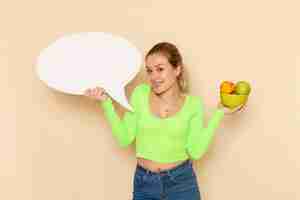 Foto grátis vista frontal jovem linda mulher de camisa verde segurando o prato cheio de frutas com placa branca na parede creme vitamina modelo de frutas mulher