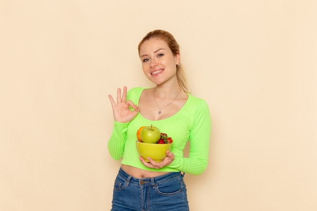 Vista frontal jovem linda mulher de camisa verde segurando o prato com frutas sorrindo na luz creme piso fruta modelo mulher pose senhora