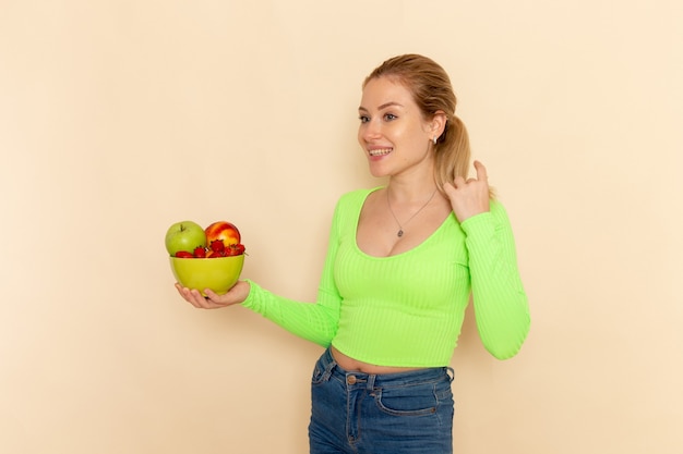 Vista frontal jovem linda mulher de camisa verde segurando um prato cheio de frutas na parede de creme claro modelo de frutas mulher cor de vitamina alimentar