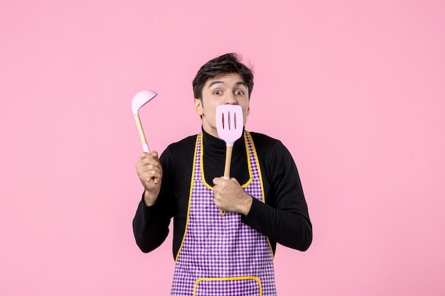 Vista frontal jovem macho na capa segurando colheres no fundo rosa profissão cor refeição cozinhar comida de trabalho horizontal massa uniforme