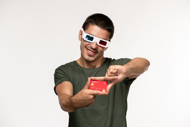 Vista frontal jovem masculino com camiseta verde segurando o cartão do banco e óculos de sol sorrindo na parede branca filme filmes de cinema solitário