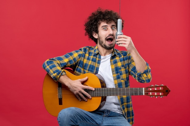 Vista frontal jovem masculino tocando violão e cantando na parede vermelha banda cantor desempenho músico concerto cor música