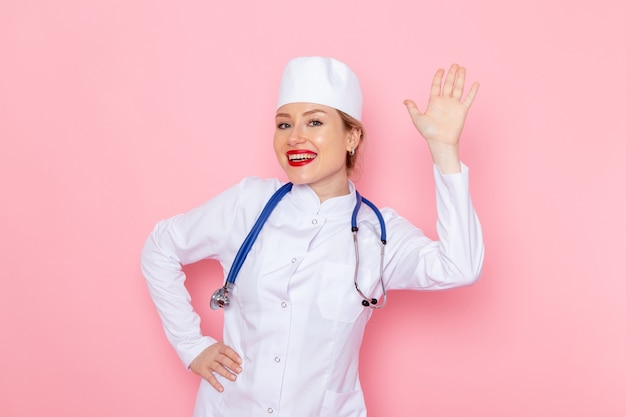 Vista frontal jovem médica em terno branco com estetoscópio azul sorrindo e posando no espaço rosa medicina