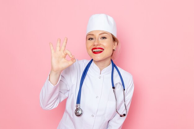 Vista frontal jovem médica em terno branco com estetoscópio azul sorrindo, posando no espaço rosa medicina médico hospitalar trabalho feminino