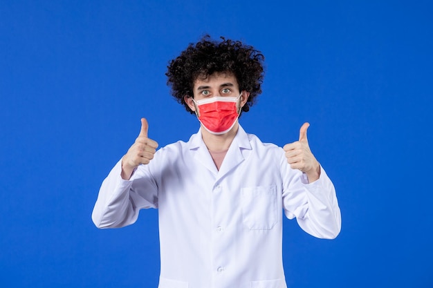 Vista frontal jovem médico em traje médico com máscara vermelha na droga de vírus de fundo azul covid- saúde da vacina hospitalar pandêmica