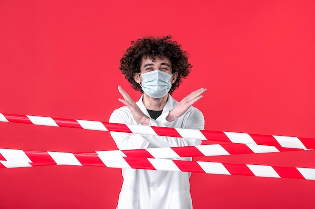Vista frontal jovem médico masculino em uniforme médico e máscara em fundo vermelho droga de isolamento covid perigo aviso quarentena de saúde